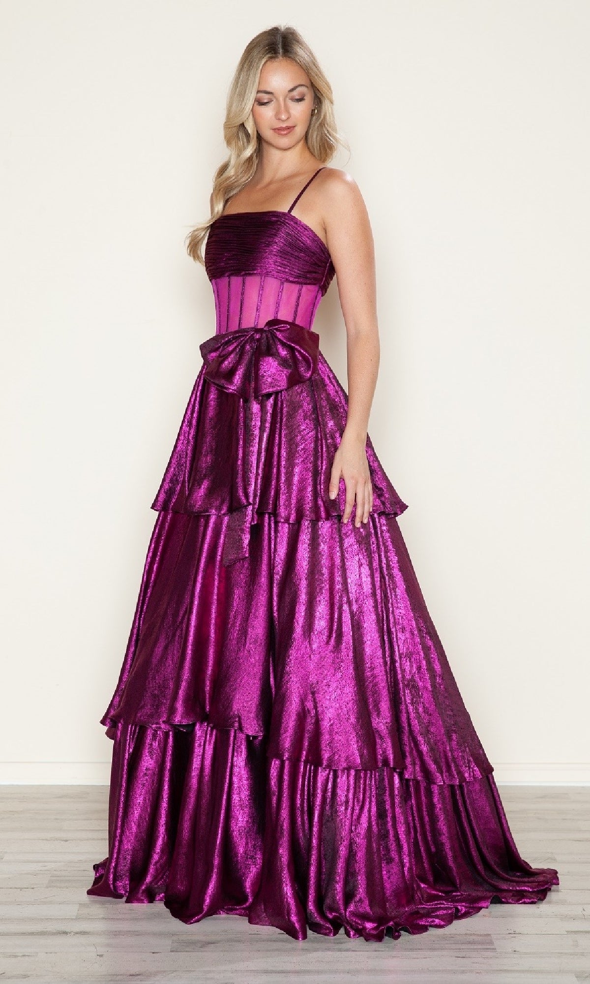 A woman poses against a plain light-colored wall, wearing the Poly USA 9718 long prom dress featuring shiny metallic fabric, spaghetti straps, a tiered A-line skirt, and a large bow at the waist.